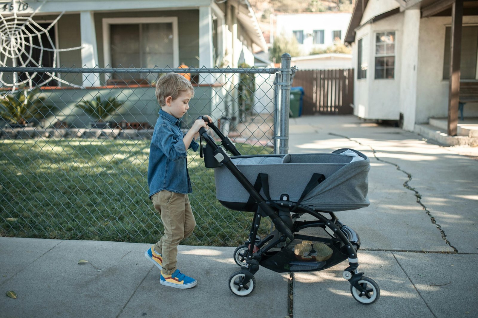 A young boy pushing a baby stroller along a suburban sidewalk on a sunny day.