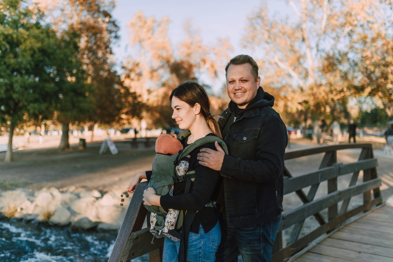 A joyful couple with a baby in a carrier at a scenic park bridge in fall.